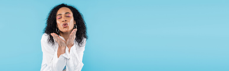 brunette african american woman with closed eyes blowing air kiss isolated on blue, banner.