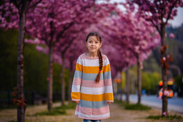 Little preschooler girl standing in striped dress before of blooming pink sakura alley