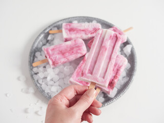 Hand holding the stick of a homemade yogurt and strawberry ice cream popsicle. In the background others on ice in a round silver tray.