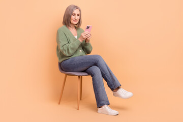 Fototapeta na wymiar Full size photo of good mood retired person wear knit pullover look at smartphone sitting on chair isolated on pastel color background