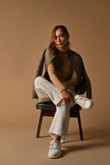 Full body portrait of Young Asian woman in cross legged pose on chair. taken at the studio on a brown background.