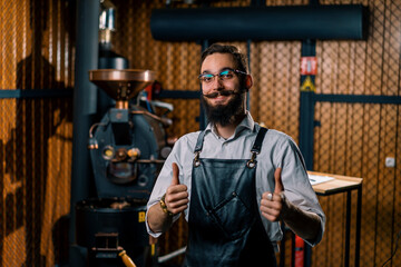 Fototapeta na wymiar satisfied handsome guy in apron and glasses stands in coffee production near machine for roasting coffee beans