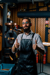Fototapeta na wymiar satisfied handsome guy in apron and glasses stands in coffee production near machine for roasting coffee beans