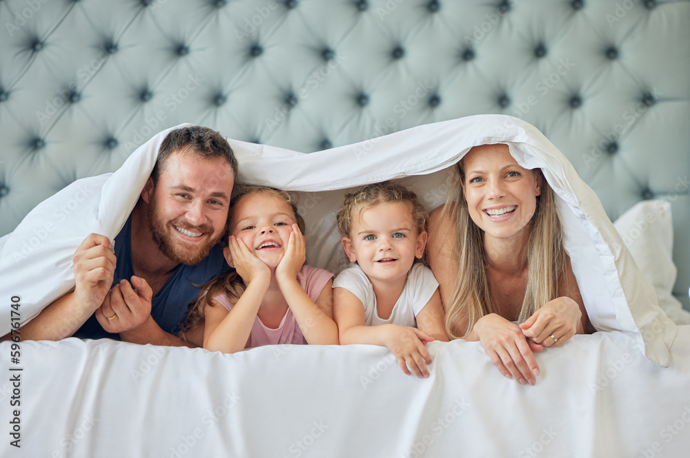 Poster A happy family in bed under a blanket at home. Portrait of smiling young parents having fun with children in the bedroom, covering with a duvet. Cute little girls playing with their mother and father