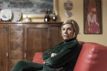 donna anziana vestita di verde è seduta nel divano di casa sua mentre guarda preoccupata - obrazy, fototapety, plakaty