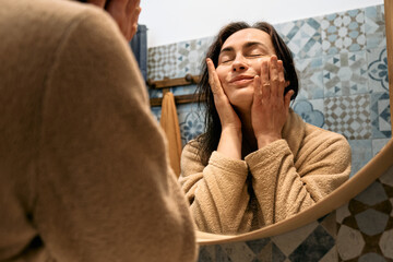 Middle-aged woman in bathrobe looking at mirror in bathroom and touching her face, applying...
