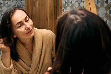 Happy middle-aged brunette woman in bathrobe looking at mirror in bathroom, smiling and touching her wet hair, applying hydrating hair cosmetics before drying it.