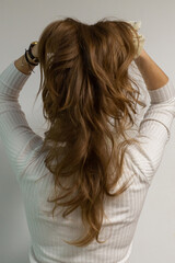 Young woman holding her natural beautiful long wavy hair from behind, model in a white blouse on a white wall, photo in a movement