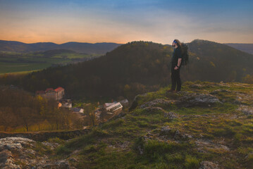 Obraz premium A lone longhaired man with a backpack on his back is standing on a sunny rocky lookout above the town of Štramberk in the Podbeskydí Nature Park.