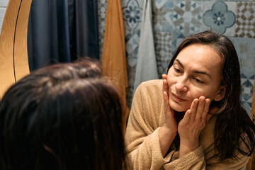 Middle-aged woman in bathrobe looking at mirror in bathroom and touching her face, applying...