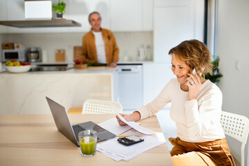 Happy senior woman talking on smartphone and checking bills when husband preparing dinner at...