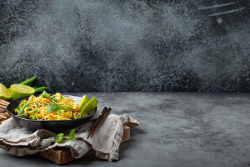 Asian vegetarian noodles with vegetables and lime in black rustic ceramic bowl, wooden chopsticks on cutting board angle view on stone background. Cooking noodles concept, copy space