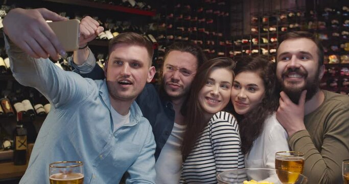 Caucasian male and female friends taking selfie photo with phone camera in bar when watching sport game. Men and women making photos on smartphone and smiling in pub. Football fans watch TV with beer.