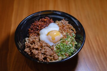 A bowl of Chili Pan Mee containing Half-Cooked Egg, Anchovy Fish, Dried Chili, and Chicken Meat on a wooden table