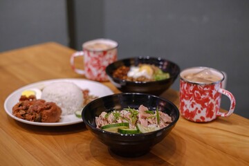 A set of Malaysian cuisine containing Steamed Chicken Rice, Nasi Lemak, Chili Pan Mee, and two mugs of Thai Tea on a wooden table with blurred background