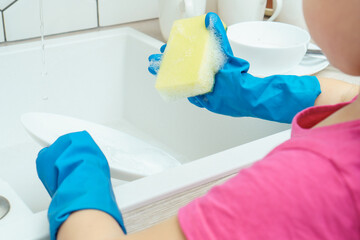 Cropped photo of little preteen girl wearing pink T-shirt, big blue rubber gloves, standing near sink, holding yellow soapy sponge, washing dishes in kitchen. Housework, housekeeping, dishwashing.