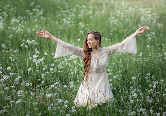 Naklejka na ściany i meble A beautiful young woman in the green grass and dandelions. Enjoy nature. A woman in a dress on a spring lawn. Freedom without allergies