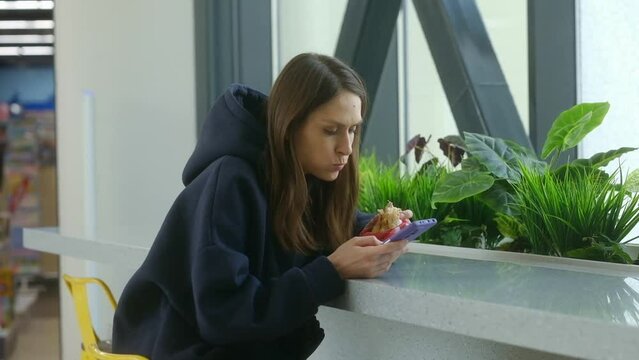 Brunette girl 20s in hoodie eating burger and serf internet on your smartphone in city cafe or city mall durning lunch. Junk unhealthy food. Concept of meat nutrition. Fast food concept.