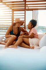 Cheerful female friends talk while relaxing on deck chair during summer day.