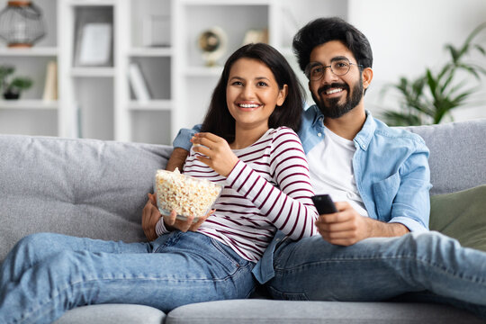 Happy multicultural couple watching TV at home, eating popcorn
