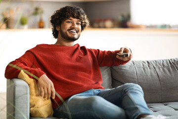 Weekend Pastime. Cheerful Indian Guy Watching Tv At Home, Holding Remote Controller