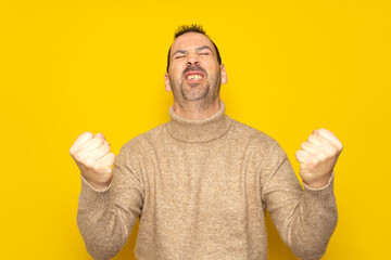 Bearded Hispanic man in his 40s wearing a beige turtleneck energetically celebrating a big win with raised fists, isolated on yellow studio background.