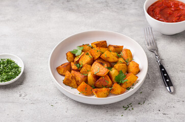 Traditional Spanish potato, patatas bravas with smoked paprika, spicy tomato sauce and parsley in a white bowl on gray stone background, top view. Delicious homemade food