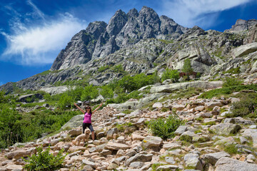 Fototapeta na wymiar Hiking trail at the beautiful mountain Restonica.View of the peaks of the Restonica mountains and a hiking trail where a young woman is walking with a dog, Lac du Melu, Corsica island, France