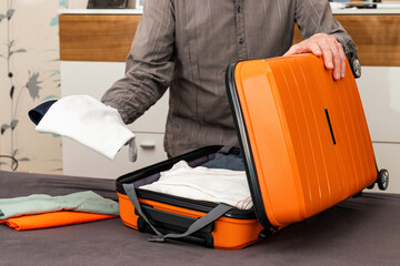 A man folds things in an orange-colored travel suitcase on wheels. Travel preparation.