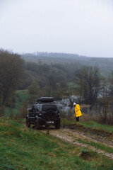A woman in a yellow jacket next to an off-road vehicle. Overcast cold spring day, landscape with river and rocky shores. A car for tourism and extreme adventures. Vertical photo