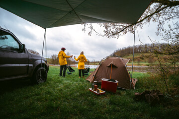 The couple is celebrating. They clink beer cans. Man and woman on vacation, traveling on an off-road vehicle. Camping equipment, tent and chairs. River view.