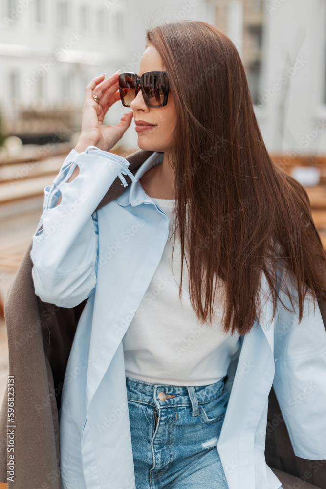 Poster Fashionable beautiful young caucasian woman in fashionable casual clothes with a coat, shirt and jeans wears with sunglasses on the street near a wooden fence