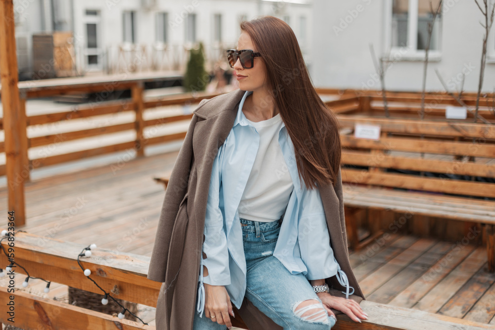 Sticker Beautiful fashionable girl with vintage sunglasses in a trendy fashion casual outfit with a stylish coat, shirt and jeans sits and rest on a wooden fence on the street