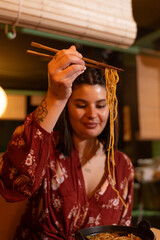 vertical shot brunette girl is dining at a Japanese restaurant. She is dressed in Japanese print clothes. She uses her chopsticks to eat yakisoba noodles.