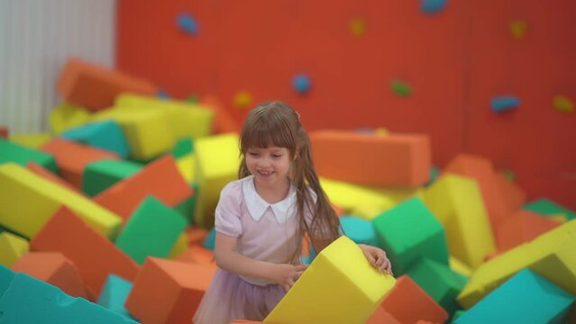 Little happy girl play and have fun in a dry pool with soft multicolored paralon cubes in a children's entertainment center and smiles in the playroom.