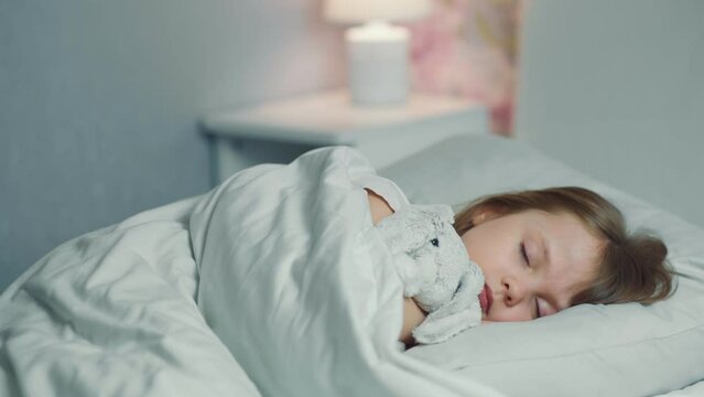 Cute funny little girl with brown hair sleeping in the white bed with favorite fluffy toy bunny. Woman hand covers with blanket caressing lying daughter. Good night. Bedtime rest. Carefree childhood