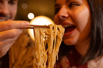 the boy holds the noodles in the bowl with his chopsticks to share them with his girlfriend, they...