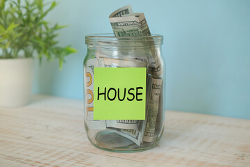 Glass jar with money and label HOUSE on table against blurred background.