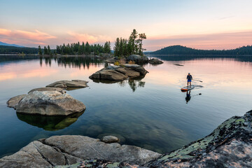 Woman paddling on tranquil Payette Lake toward rocky islands at sunset. - obrazy, fototapety, plakaty