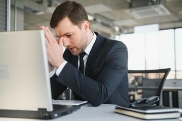 Young stressed handsome businessman working at desk in modern office shouting at laptop screen and being angry about financial situation, jealous of rival capabilities, unable to meet client needs