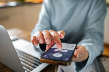 Young adult woman using a banking app on her phone while working from home