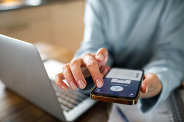 Young adult woman using a banking app on her phone while working from home