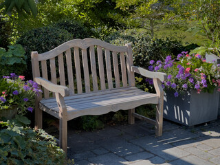 Ornate wooden bench sits empty on a stone patio in a flowering garden.