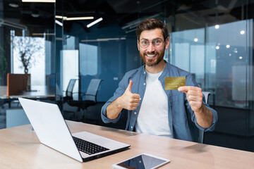 Fototapeta na wymiar Bank credit card advertising. A young man sits in the office at the table and holds a golden money card, offers to buy, shows a super hand to the camera.