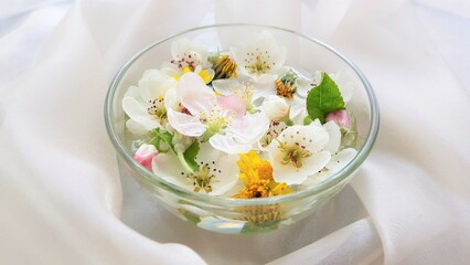 Yellow dandelions, white blooming flowers, green leaves floating in a bowl of water on a white thin material.