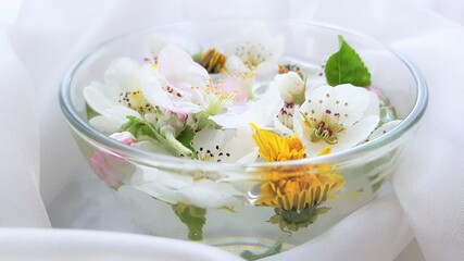 Yellow dandelions, white blooming flowers, green leaves floating in a bowl of water on a white material.