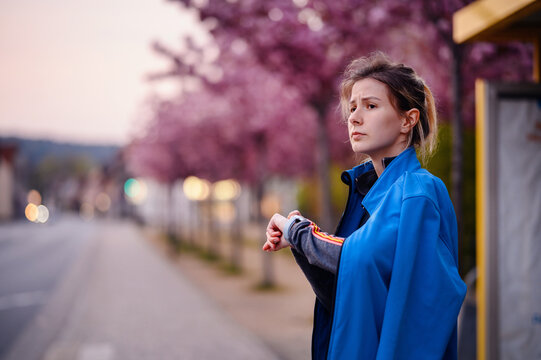 Young girl in blue jacket and jeans standing on bus stop with sakura alley looking on watch on hand