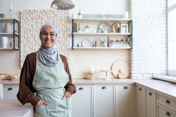Portrait of Arab young housewife woman in hijab standing in kitchen near table in apron, holding...