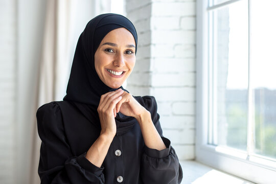Portrait of a young beautiful woman in a black burqa, hijab standing by the window, smiling at the camera and holding her hands togehter near her face.