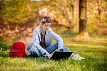 Young girl in spring outwear working on laptop on green grass in parks